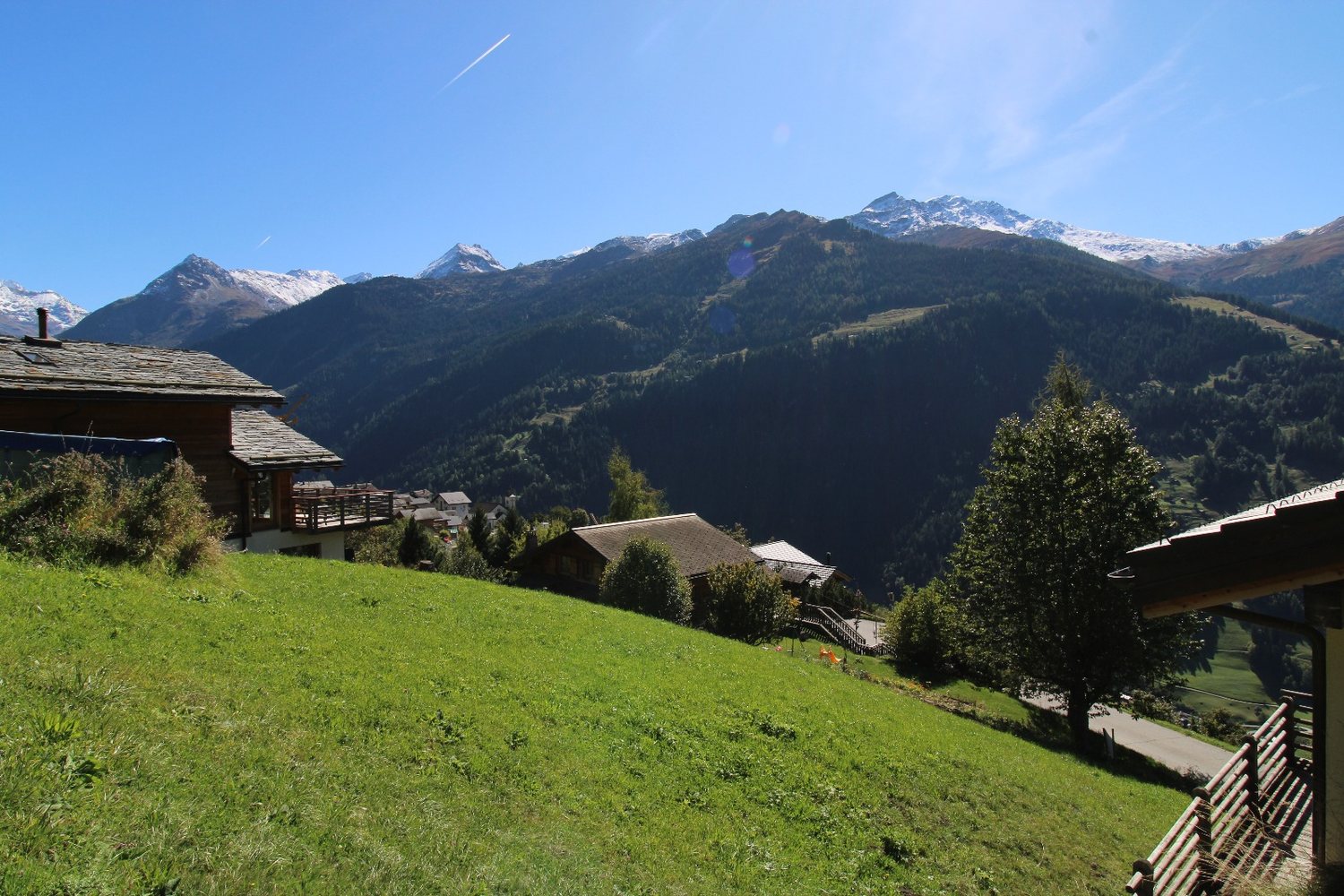 Le pré en pente douce, chalets à gauche et à droite, vallée boisée et crêtes au fond.