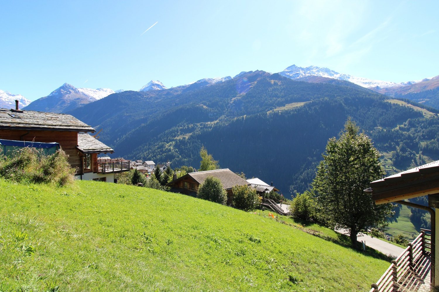 Le pré de la parcelle entre deux chalets en madrier, avec les sommets enneigés du Val de Bagnes en arrière-plan.