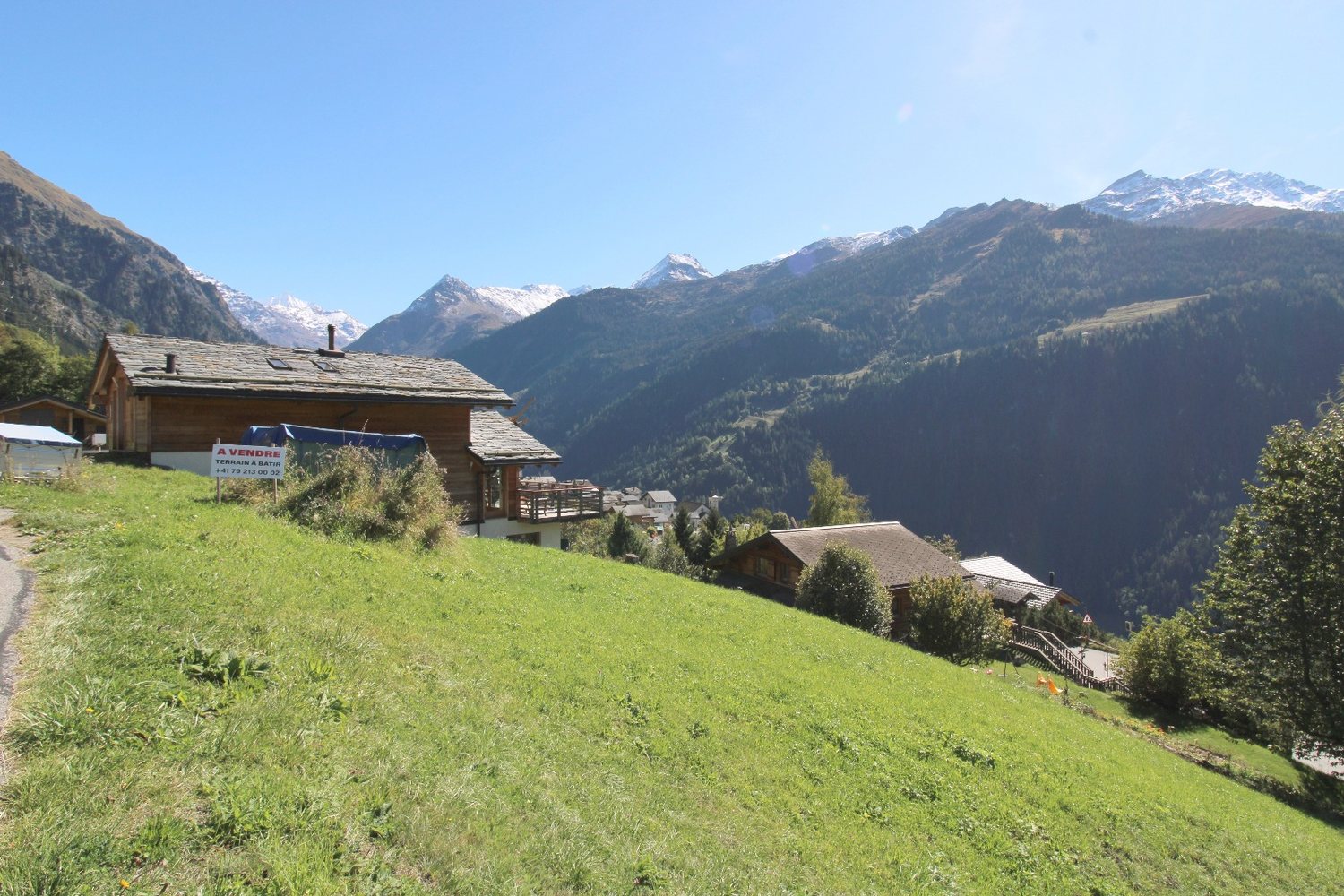 Le pré vu depuis le chemin, chalets en madrier à gauche, vallée et sommets en arrière-plan.