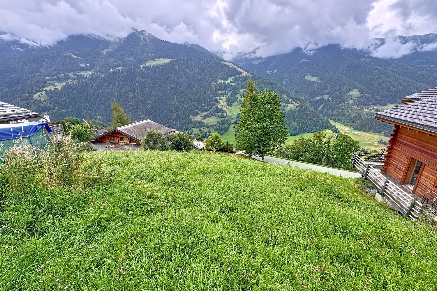 Vue élargie depuis le haut de la parcelle : le pré, les chalets voisins, la vallée de Bagnes et les crêtes sous un ciel de nuages.